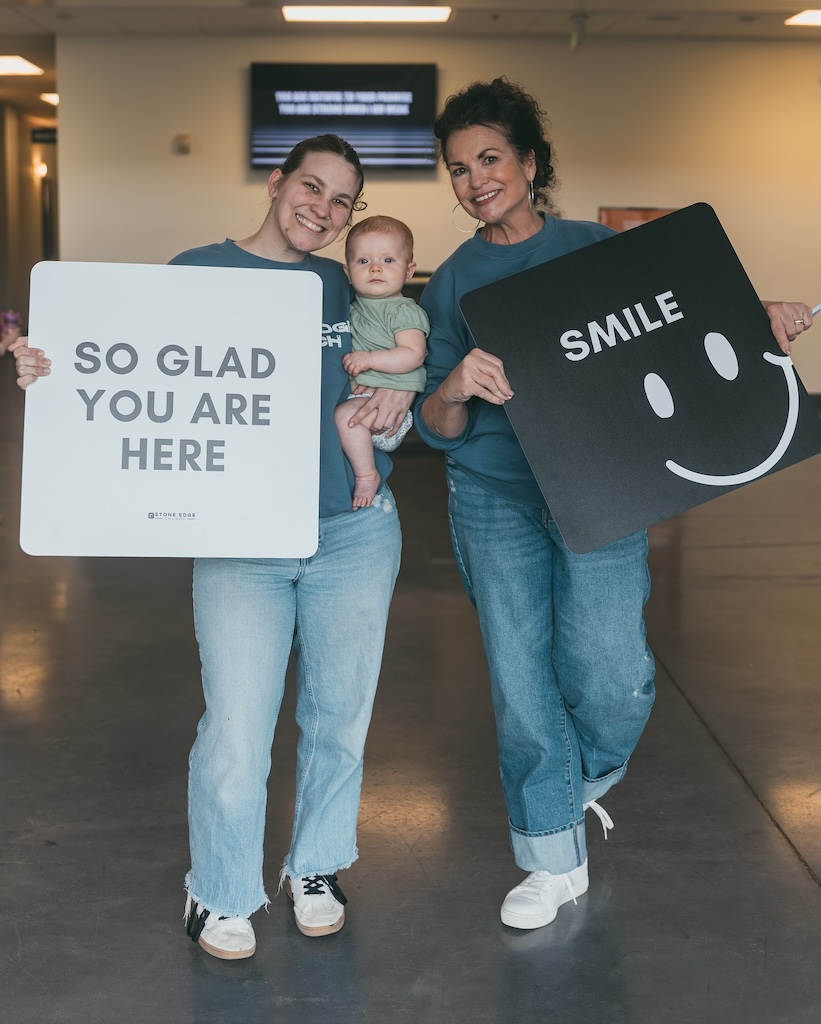Two women in Stone Edge Church shirts holding "So glad you are here" and "Smile" welcome signs with a baby